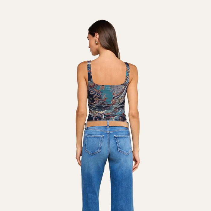 A woman with straight brown hair stands facing away, wearing the Rasha Shirred Mesh Tank—a versatile women’s top with a square neckline—paired with blue jeans and a visible tan belt, set against a plain light background.