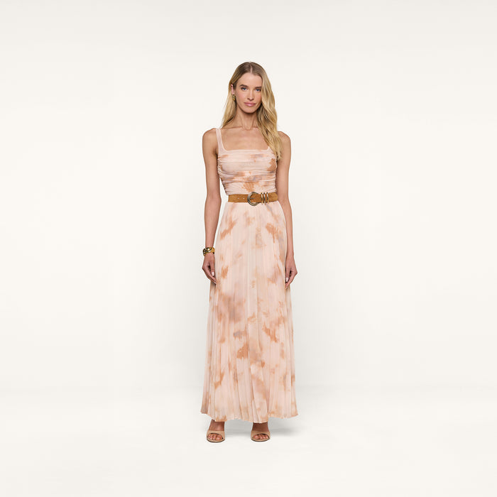 A woman stands against a plain white background, wearing the Rasha Shirred Mesh Tank—a sleeveless, floor-length dress with an abstract peach and white tie-dye print. She accessorizes with a tan belt, brown sandals, and a bracelet. Her long blonde hair frames her neutral expression.