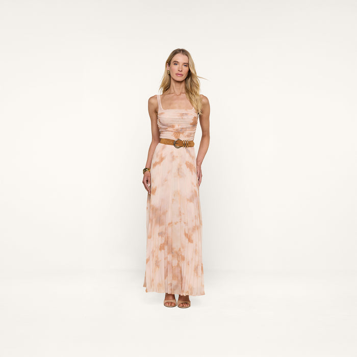 A woman stands against a white background wearing the Rasha Shirred Mesh Tank styled as a long, sleeveless dress with an abstract light pink and beige print, paired with a belt and heeled sandals.