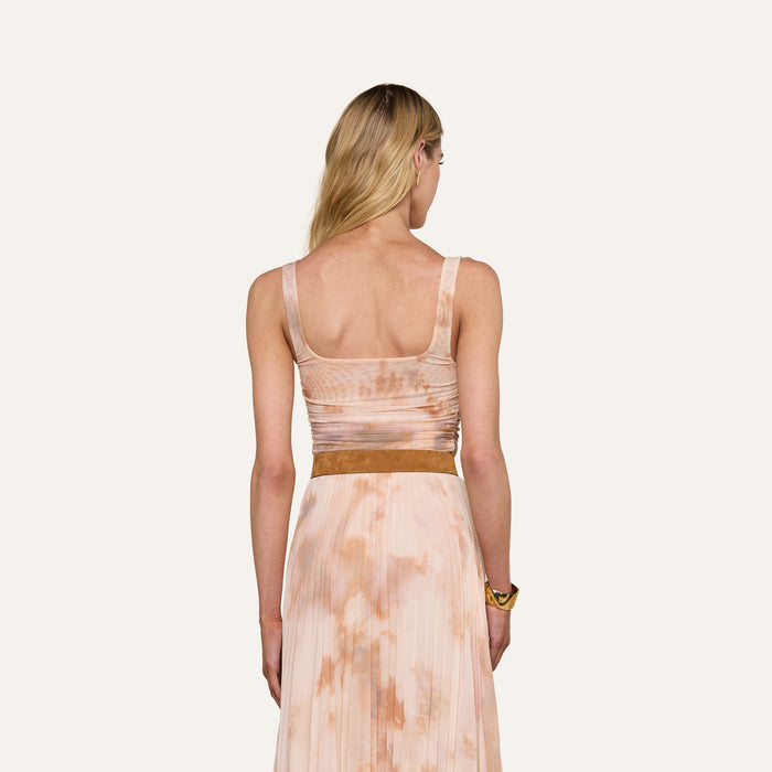 A woman with long blonde hair is shown from behind, wearing the Rasha Shirred Mesh Tank, a sleeveless top in a light pink and tan abstract print paired with a brown belt. She has a gold bracelet on her left wrist. The background is plain and light-colored.