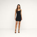 A woman stands in a plain room wearing the fitted, sleeveless Raye Slip Lining Dress crafted from stretch jersey fabric, paired with black high-heeled sandals. She has long brown hair and a neutral expression as she faces the camera.