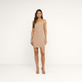A woman stands against a plain white background wearing the Raye Slip Lining Dress—a sleeveless, beige mini dress crafted from soft poly georgette fabric with adjustable straps and a slip lining. She pairs it with beige high-heeled sandals and has long brown hair and a neutral expression.