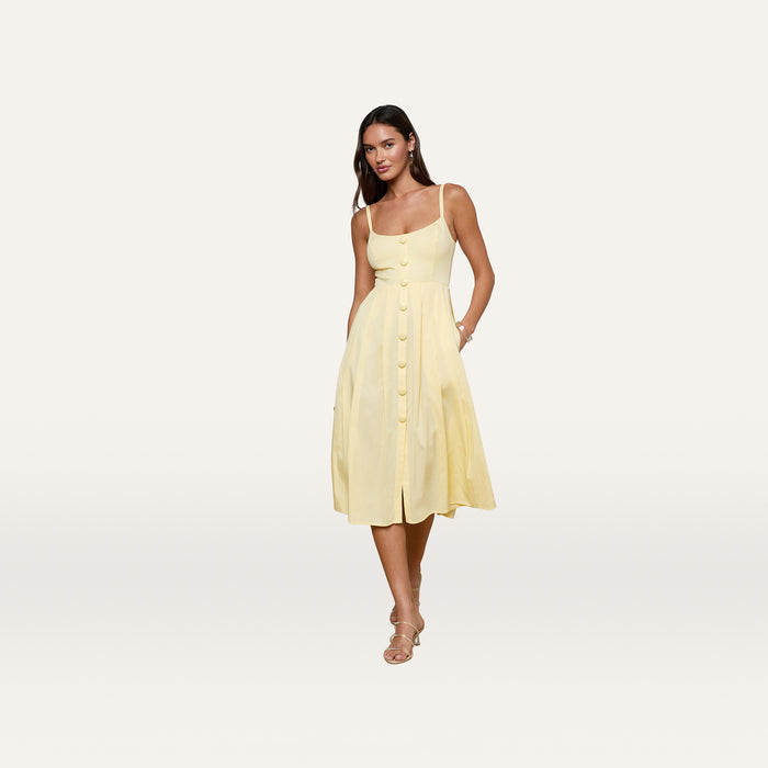 Wearing the Reanne Midi Dress—a sleeveless, light yellow poplin midi with a smocked back and large front buttons—a woman pairs it with beige sandals, posing against a plain white background.