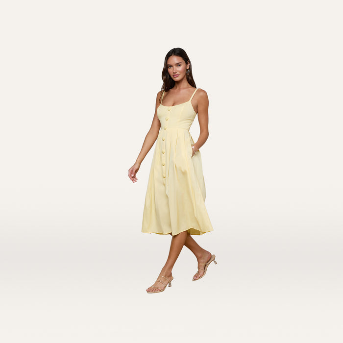 A woman stands against a plain white background wearing the Reanne Midi Dress, a sleeveless, light yellow poplin midi dress featuring a fitted bodice, smocked back, and flared skirt, paired with beige heeled sandals.