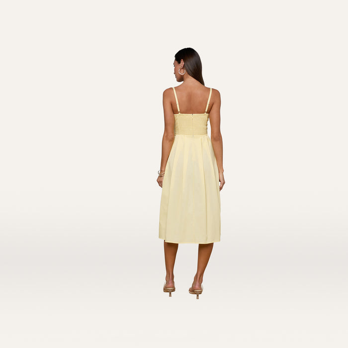 A woman stands with her back to the camera on a plain white background, wearing the Reanne Midi Dress in light yellow—a sleeveless poplin dress featuring a smocked back—paired with high-heeled shoes.