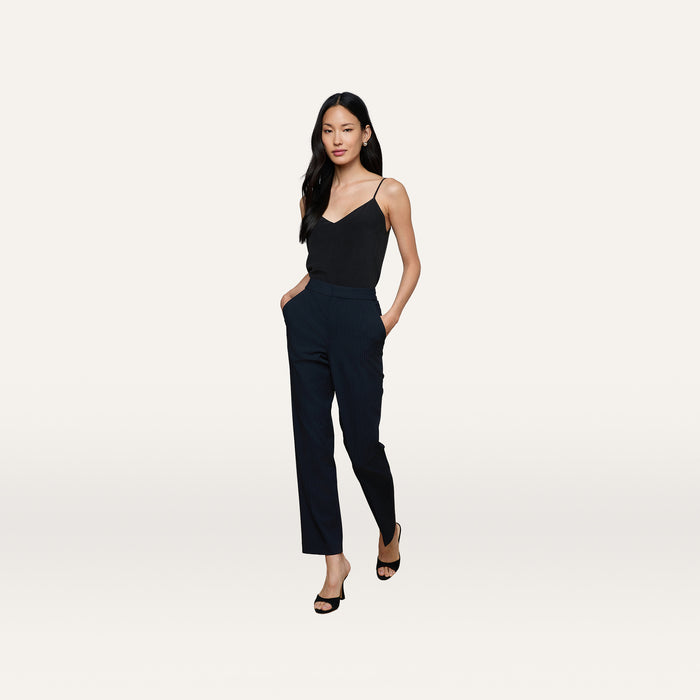 A woman stands with her hands in her pockets, wearing a black sleeveless top, the high-rise Rebel Pinstriped Trouser in navy, and black heels against a plain white background.