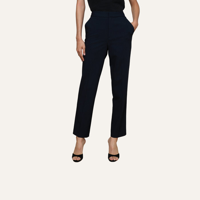Wearing the Rebel Pinstriped Trouser in black high rise, paired with black open-toe heels and hands casually tucked into pockets, set against a plain light background. Upper body is partially out of frame.