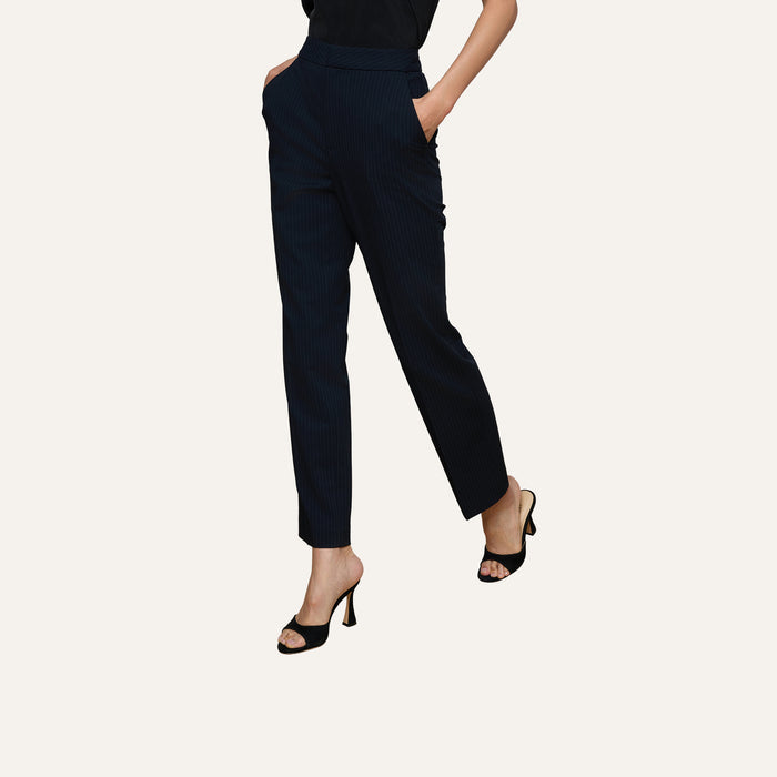 A person wearing the Rebel Pinstriped Trouser—a high-rise, straight-leg style with navy pinstripes—paired with a black top and black high-heeled sandals stands with hands in pockets against a plain background.