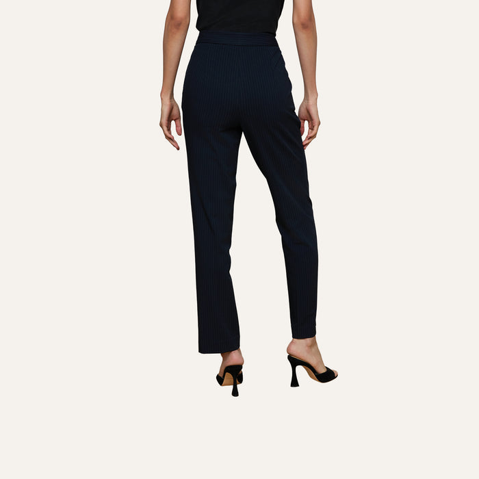 A person wearing the Rebel Pinstriped Trouser, featuring a high-rise, straight-leg silhouette with navy pinstripes, and black heeled shoes stands with their back to the camera against a plain light background.