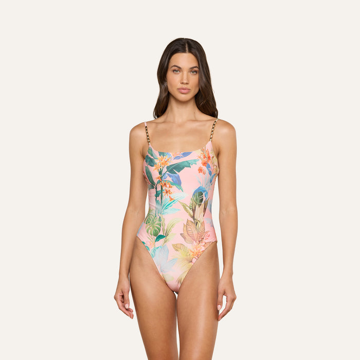 A woman models the Remi One-Piece Swimsuit, featuring a high-leg cut, chain straps, and a tropical floral and palm leaf print, standing against a plain off-white background.