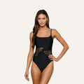 A woman exudes poolside glamour in the Remi One-Piece Swimsuit—a black maillot adorned with butterfly cutout designs on the sides—standing against a plain, light background with one hand on her hip.