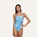 A woman models the Remi One-Piece Swimsuit, featuring a white baroque print with blue patterns, as she poses with one hand on her hip against a plain light background.