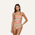 A woman with long brown hair wears the Remi One-Piece Swimsuit, featuring watercolor stripes, thin straps, and a high-leg cut. She stands against a plain light gray background, smiling softly at the camera.