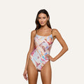 A woman models the Remi One-Piece Swimsuit, featuring UPF 50 fabric with travel-themed postcard prints and chain hardware straps, posing with one hand on her hip against a plain, light background.