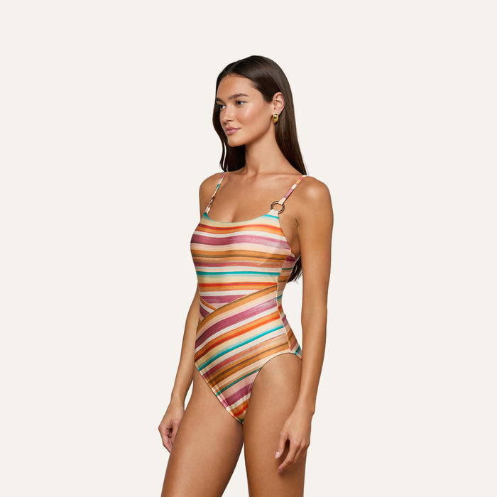 A woman with long brown hair stands against a plain background, wearing the Remi One-Piece Swimsuit featuring colorful watercolor stripes, thin shoulder straps, and a high-leg cut. She accessorizes with gold hoop earrings and faces slightly to the side.