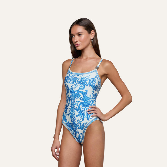 A woman models the Remi One-Piece Swimsuit, featuring a blue and white baroque print and thin straps, as she stands with one hand on her hip against a plain light background.