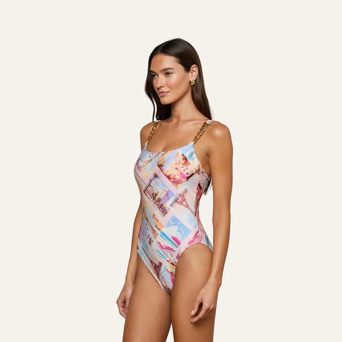 A woman wears the Remi One-Piece Swimsuit, which features postcard print and gold chain hardware straps, as she stands against a light background looking slightly to the side with a neutral expression.
