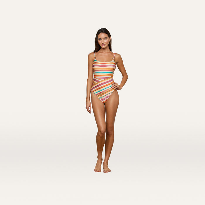 A woman stands barefoot on a wooden floor in the Remi One-Piece Swimsuit, featuring watercolor stripes, cutouts, and a high-leg cut. She faces forward with a slight smile, arms relaxed at her sides against a plain background.