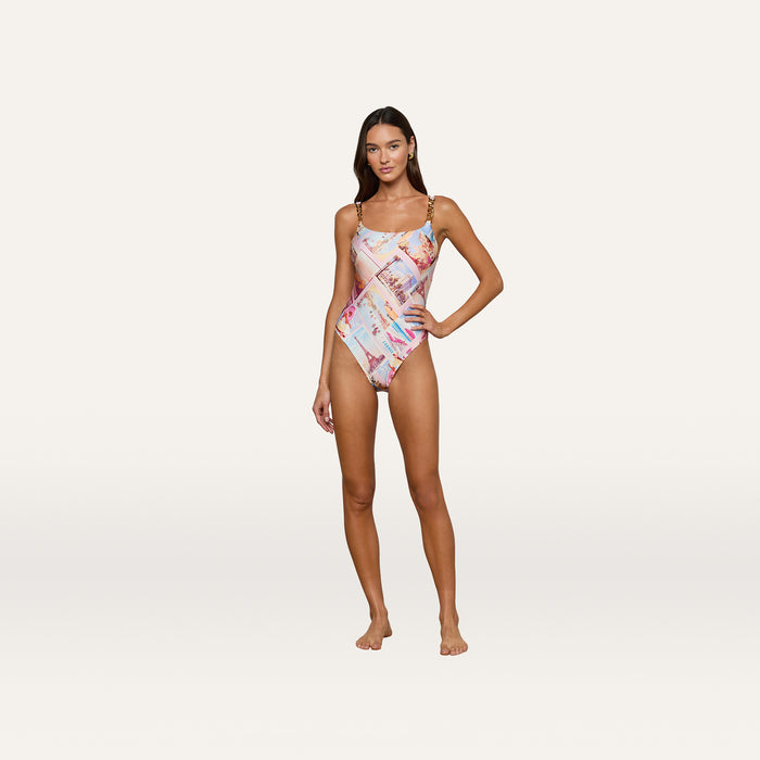 A woman poses barefoot against a plain, light background, wearing the Remi One-Piece Swimsuit featuring a colorful geometric postcard print.
