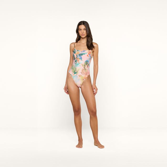 A woman stands barefoot against a white background, modeling the Remi One-Piece Swimsuit with thin straps and a high-leg silhouette. Her long hair is worn loose, her expression neutral, and her arms rest naturally at her sides.