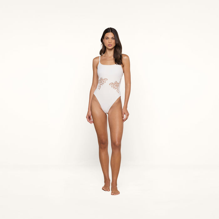 A woman stands barefoot against a plain background, wearing the Remi One-Piece Swimsuit, a white high-leg cut one-piece with cutout lace details on the sides.