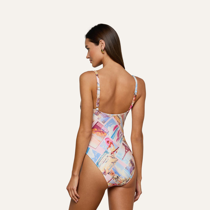 A woman with long brown hair is shown from the back, wearing the Remi One-Piece Swimsuit. This colorful postcard print maillot features a collage pattern, thin straps, a low back, and UPF 50 protection. The background is plain and light-colored.