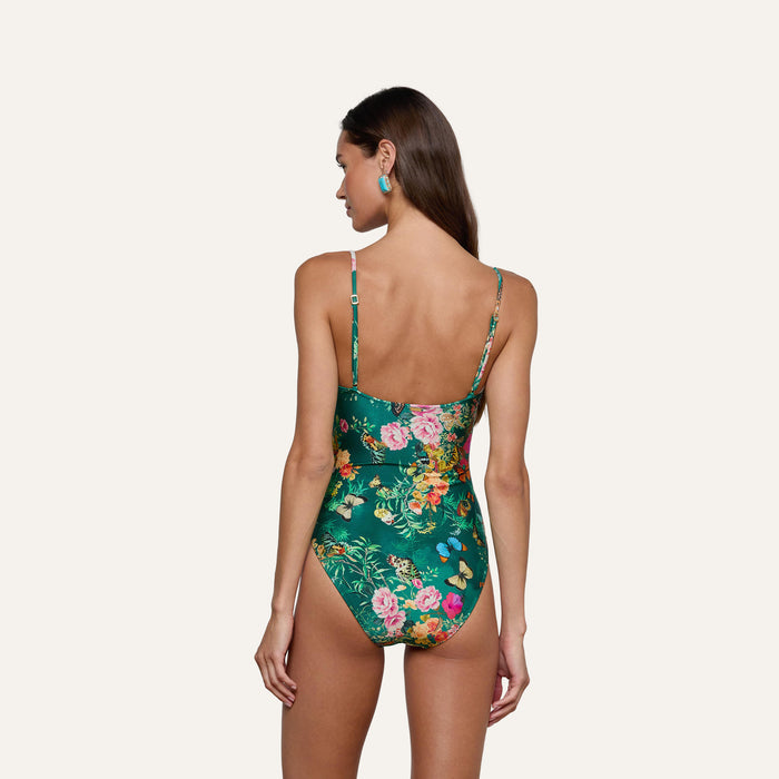 A woman with long brown hair is seen from behind, wearing the Remi One-Piece Swimsuit in a green floral-butterfly print with adjustable straps, standing against a plain light background.