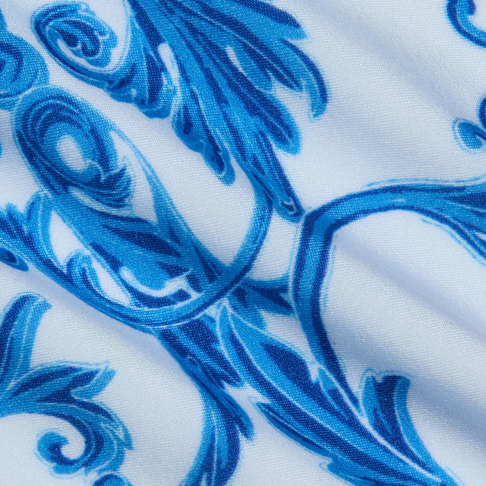 Close-up of the Remi One-Piece Swimsuit, showcasing its white fabric adorned with a blue ornate floral pattern. The design features swirling and leaf-like baroque-inspired motifs, while subtle wrinkles in the fabric enhance the textured look of this decorative swimwear.