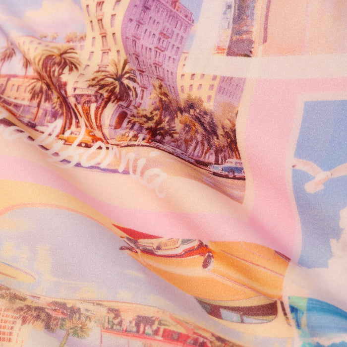 The Remi One-Piece Swimsuit features a colorful, pastel-toned fabric adorned with palm trees, buildings, vintage cars, and the word “California,” evoking a retro beach-inspired scene reminiscent of a postcard print. The softly creased texture adds to its nostalgic charm.