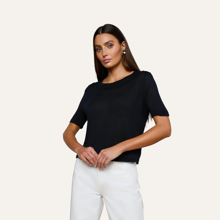 A woman with long dark hair stands confidently against a plain light background, wearing the Remy Tee—a relaxed fit, black short-sleeve top in micro modal jersey—and white pants, her hands resting at her waist as she looks at the camera.