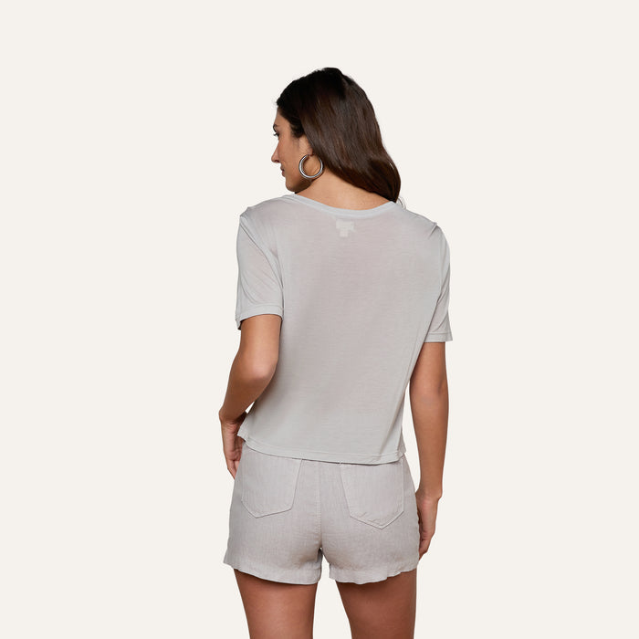 A woman with long brown hair, wearing hoop earrings and the Remy Tee in light gray micro modal jersey with matching relaxed-fit shorts, stands with her back to the camera against a plain white background.