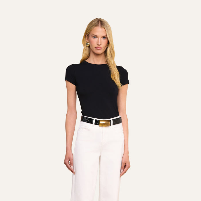 A woman with long blonde hair wears the Ressi Fitted Tee in black, paired with white pants, a black and gold belt, and gold earrings, standing against a plain light background.