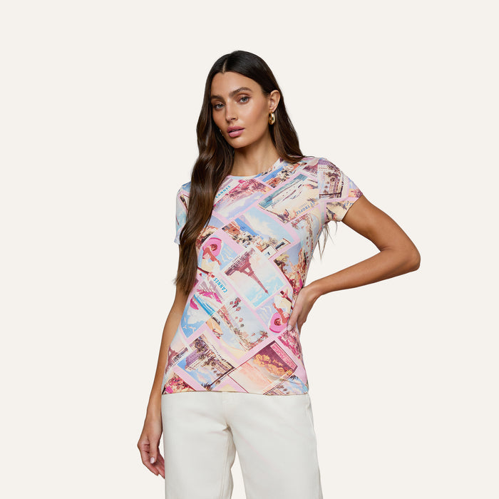 A woman with long brown hair stands confidently against a plain background, wearing the bestselling Ressi Fitted Tee in a colorful postcard print paired with white pants, one hand on her hip as she looks at the camera.
