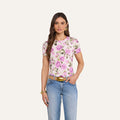 A woman stands against a light background wearing the bestselling Ressi Fitted Tee in white with a pink floral print, crafted from stretch jersey knit, paired with blue jeans, a brown belt, gold earrings, and a gold bracelet, with one hand in her pocket.