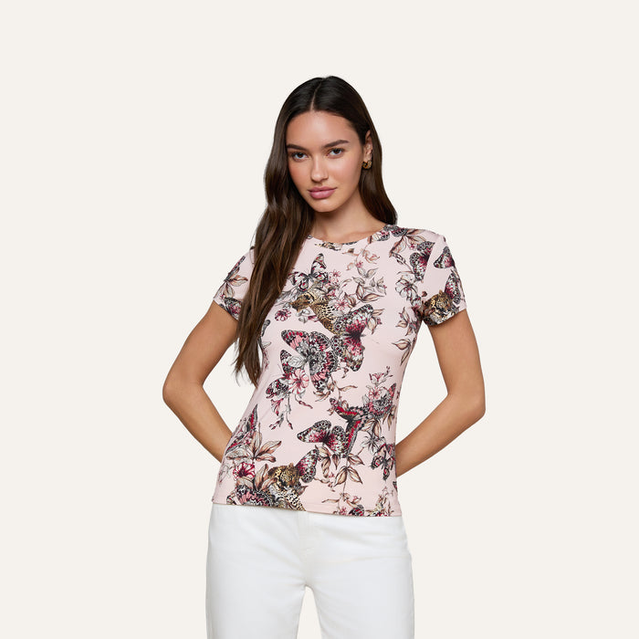 A woman stands against a plain background wearing the bestselling Ressi Fitted Tee, a light pink short-sleeve top crafted from stretch jersey knit and adorned with a vibrant butterfly jungle print, paired with white pants.