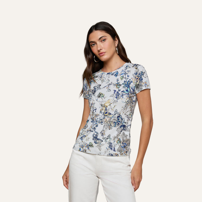 A woman with long brown hair wears the bestselling Ressi Fitted Tee, crafted from stretch jersey knit in a white floral-patterned short-sleeve design, paired with white pants. She stands against a plain light background with a neutral expression.