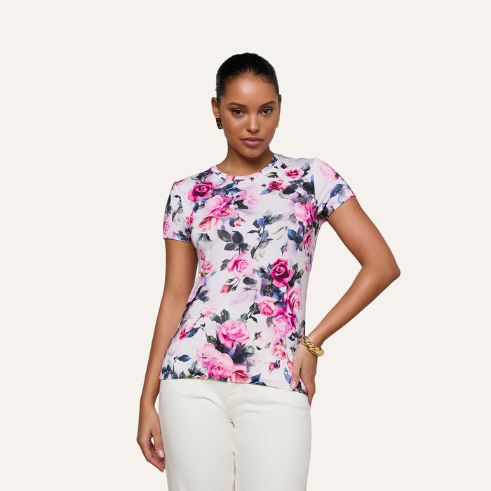 A woman poses with one hand on her hip against a plain light background, wearing the Ressi Fitted Tee—a bestselling white short-sleeved top featuring a pink rose and purple floral print—paired with white pants.