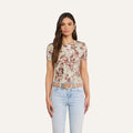 A woman stands facing forward, wearing the Ressi Fitted Tee—a slim cut floral short-sleeve top made from stretch jersey—paired with light blue jeans and a beige belt. She has long brown hair and a neutral expression, set against a plain off-white background.