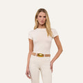 A woman with long blonde hair wears the vintage-inspired Ressi Fitted Tee in white, paired with light-colored pants and accessorized with a beige belt featuring a large gold buckle, standing against a plain light background.