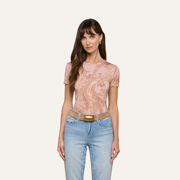 A woman with long brown hair is wearing the Ressi Fitted Tee in a mauve paisley print stretch jersey knit, paired with light blue jeans and a tan belt, standing against a plain off-white background.