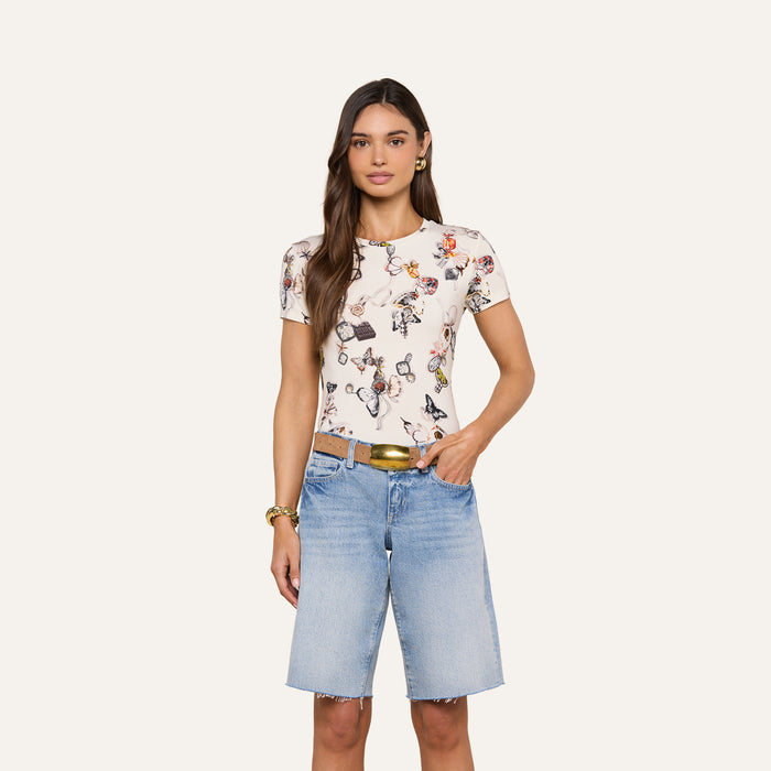 A woman stands against a plain background, wearing the bestselling Ressi Fitted Tee in printed white and slim cut light blue denim Bermuda shorts, accessorized with a gold belt and gold bracelets. She has long brown hair and a neutral expression.