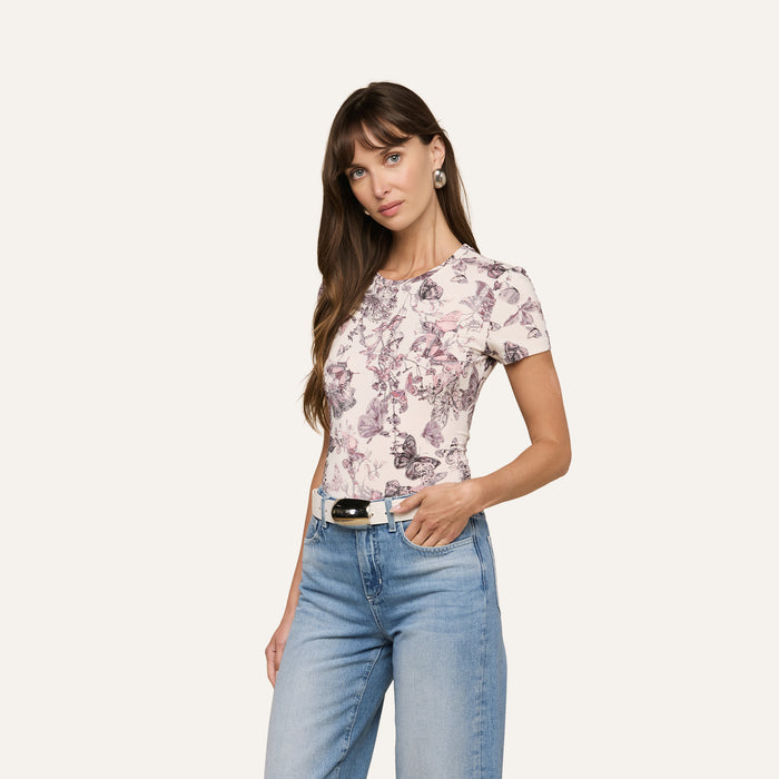A woman with long brown hair wears the bestselling Ressi Fitted Tee in a floral short-sleeve style, paired with light blue jeans, a black belt, and hoop earrings. She stands against a plain light background with one hand in her pocket.