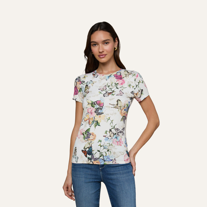 A woman wearing the Ressi Fitted Tee, showcasing a colorful butterfly garden print, paired with blue jeans, stands against a plain light background and smiles gently at the camera.