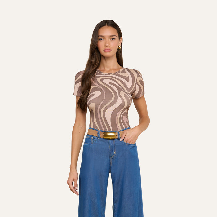 Woman with long brown hair wears the Ressi Fitted Tee in a beige and brown abstract swirl print stretch jersey knit, paired with wide-leg blue jeans, a gold buckle belt, and gold earrings, standing against an off-white background.