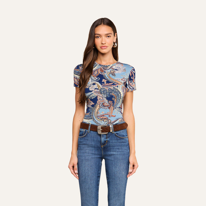 A woman with long brown hair wears the Ressi Fitted Tee in midnight blue paisley, styled with blue jeans, a brown belt, and hoop earrings as she stands against a plain light background.