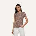 A woman stands against a light background, wearing the Ressi Fitted Tee—a slim cut brown short-sleeve shirt with white polka dots—paired with white pants, with one hand in her pocket.