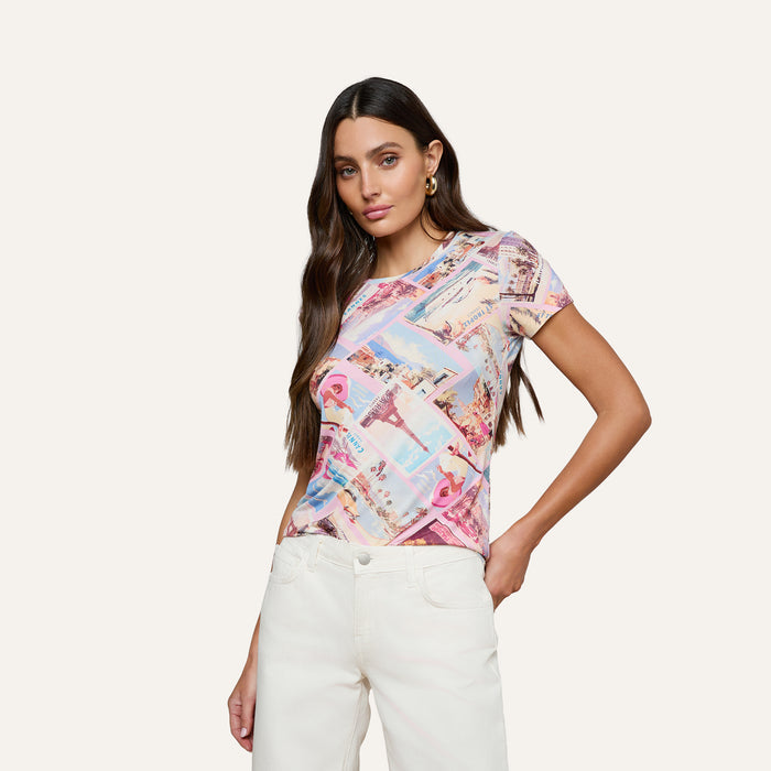 A woman with long dark hair poses against a plain light background, wearing the Ressi Fitted Tee—a colorful, patterned stretch jersey knit short-sleeve shirt—and white pants, with one hand in her pocket.
