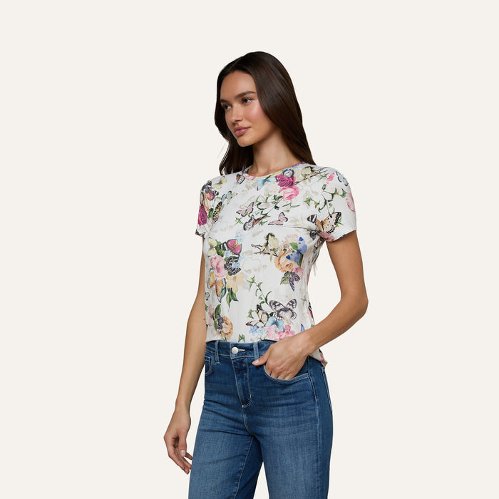 A woman stands against a plain light background wearing the Ressi Fitted Tee, a white short-sleeve shirt with a butterfly garden print, paired with blue jeans.