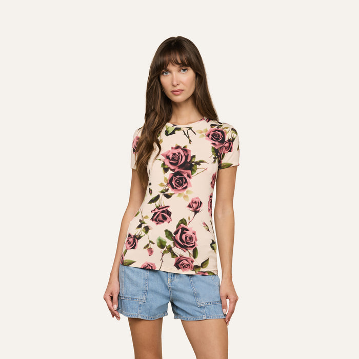 A woman wearing the Ressi Fitted Tee, a slim cut, light-colored t-shirt with a pink rose floral pattern, paired with blue denim shorts and standing against a plain, off-white background.
