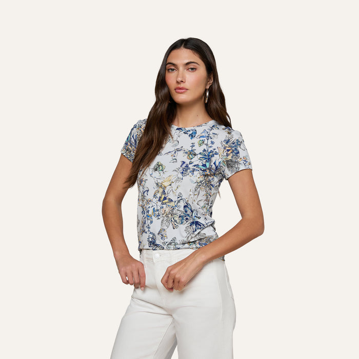 A woman with long brown hair wears the Ressi Fitted Tee—a light-colored, short-sleeve top with a blue butterfly floral print—paired with white pants, standing against a plain, light background with her hands near her waist and looking confidently at the camera.